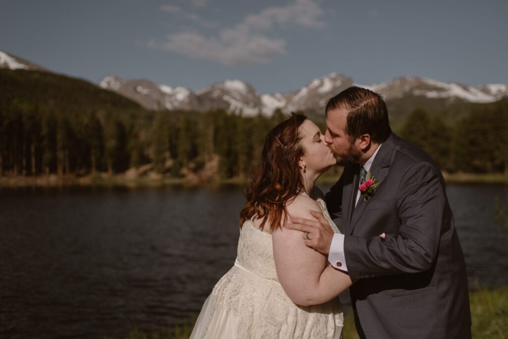bride and groom sharing first kiss at Sprague Lake with mountains and trees in distance