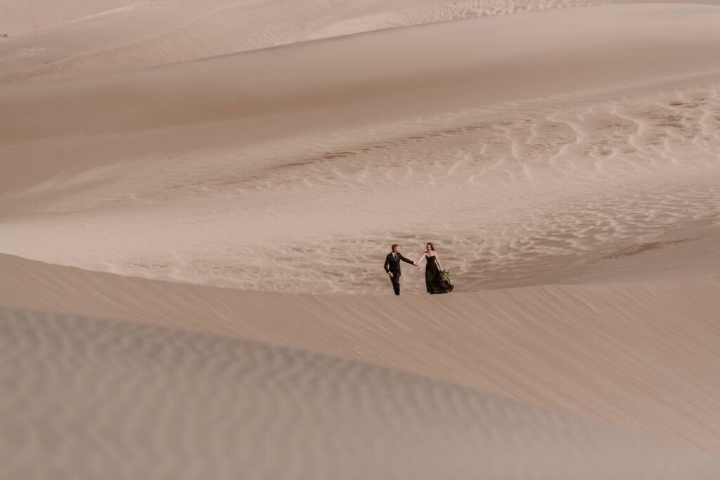 Bride and groom in all black walking across sand dunes in Colorado