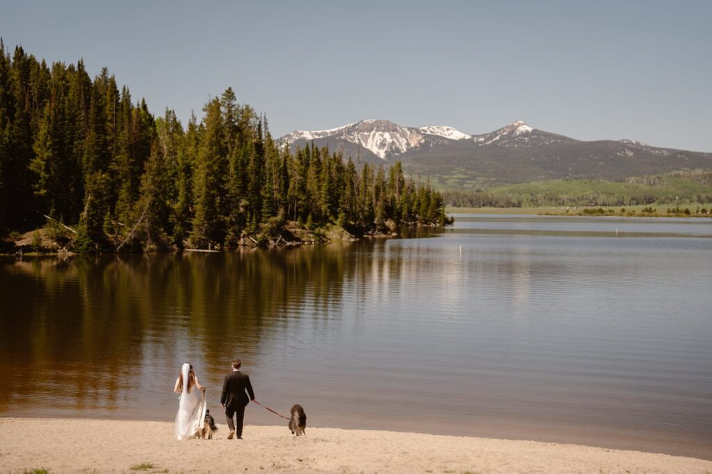 Bride, groom, and two dogs walking down to the shore of a mountain lake