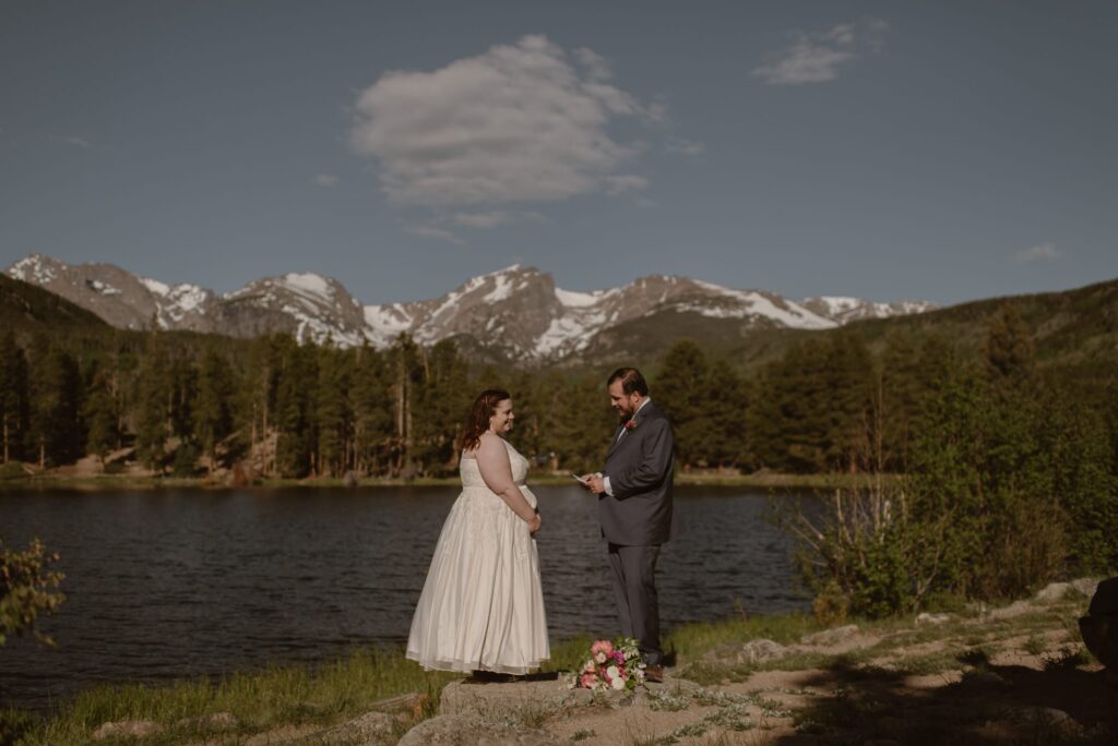 wedding couple standing on the shore of Sprague Lake in Rocky Mountain National Park with mountains in the distance
