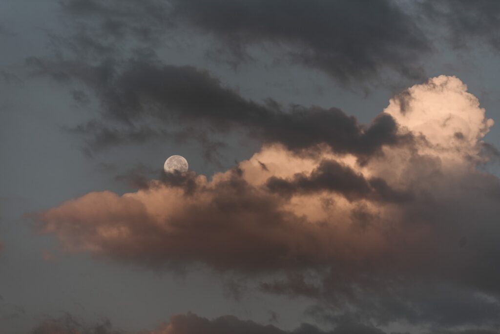 Photo of moon glowing from behind pink clouds in a blue sky
