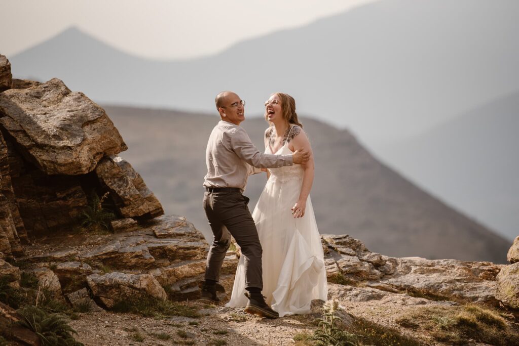 Wedding couple laughing while standing on a cliff with mountains in the distance