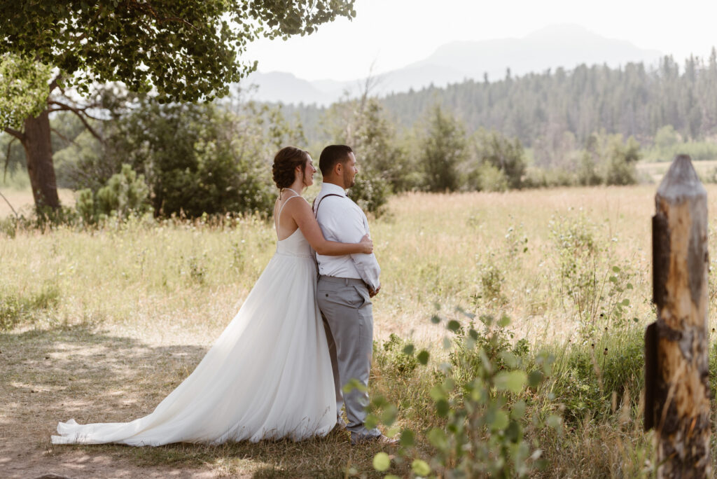 Bride hugging groom from behind at the edge of the meadow before he sees her for the first time
