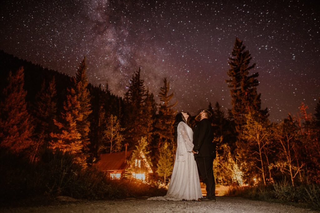 Wedding day portraits after dark in front of a glowing cabin in the woods with the milky way