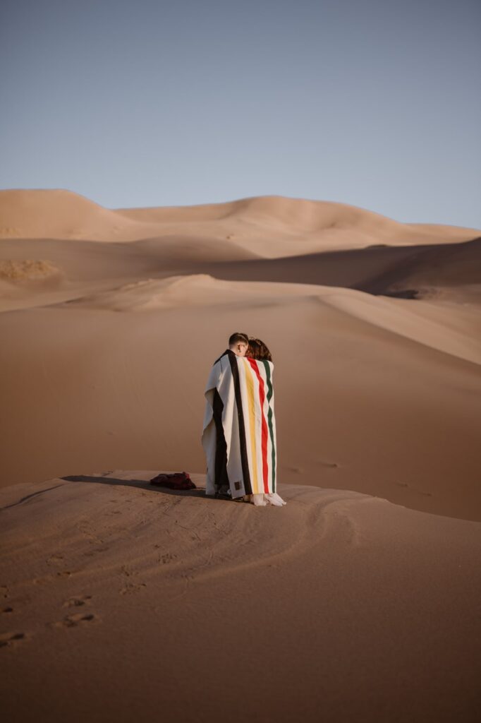 Couple hugging in a blanket on the sand dunes at sunrise