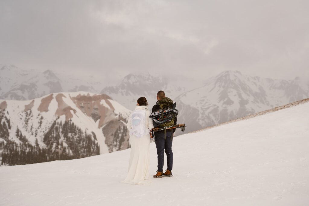 Couple with snowshoes looking out from the top of a snowy mountain on their wedding day
